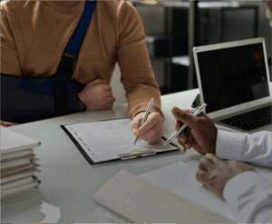 Two people sit at a desk; one has an arm in a sling, while the other points at a form on a clipboard next to a laptop and stacked documents.