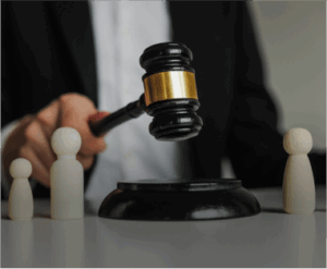 A judge in a black suit holds a gavel above a sound block, with three wooden human-shaped figures on the table.