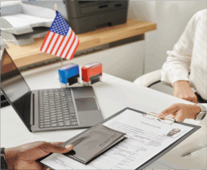 Two people at a desk exchange a clipboard with a form and a passport; an open laptop and small American flag are visible in the background.