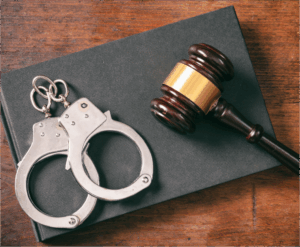 A pair of handcuffs and a wooden judge’s gavel rest on a closed black book on a wooden surface.