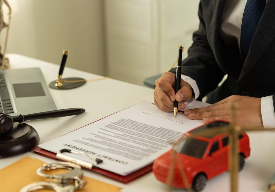 A person in a suit signs a legal document at a desk with a gavel, handcuffs, a toy car, a laptop, and legal symbols.